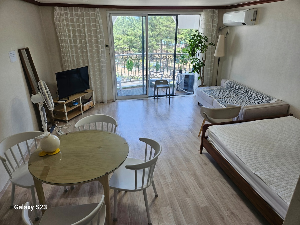 A spacious living area displays a round dining table surrounded by white chairs. A comfortable sofa is positioned next to a large glass door that opens to a balcony, providing natural light and views of greenery. A television and fan complete the inviting space.
