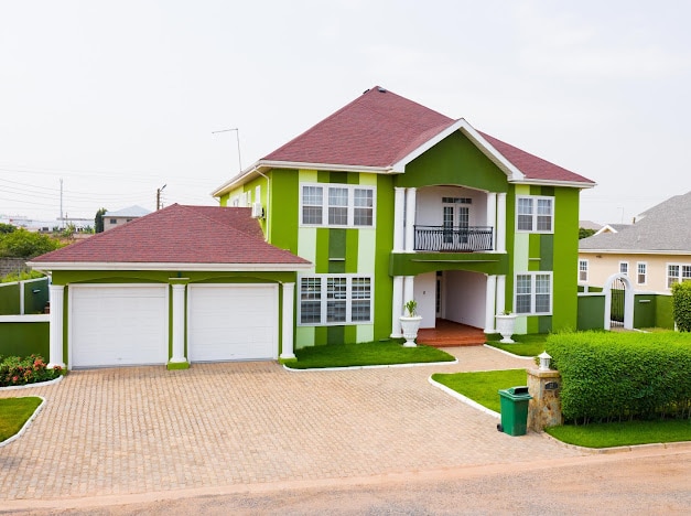 A two-story house with vibrant green and white exteriors is featured, showcasing a red tile roof and a charming balcony. The driveway leads to a double garage, with neatly trimmed hedges and flower beds accentuating the front yard.