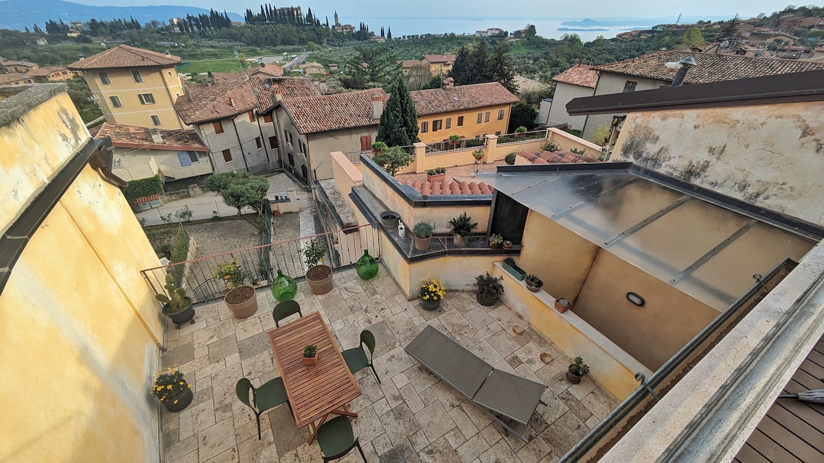 An expansive terrace is shown, featuring a wooden dining table surrounded by dark green chairs. Potted plants add greenery to the space. A sun lounger is positioned nearby, with scenic views of the surrounding hills and Lake Garda visible in the distance.