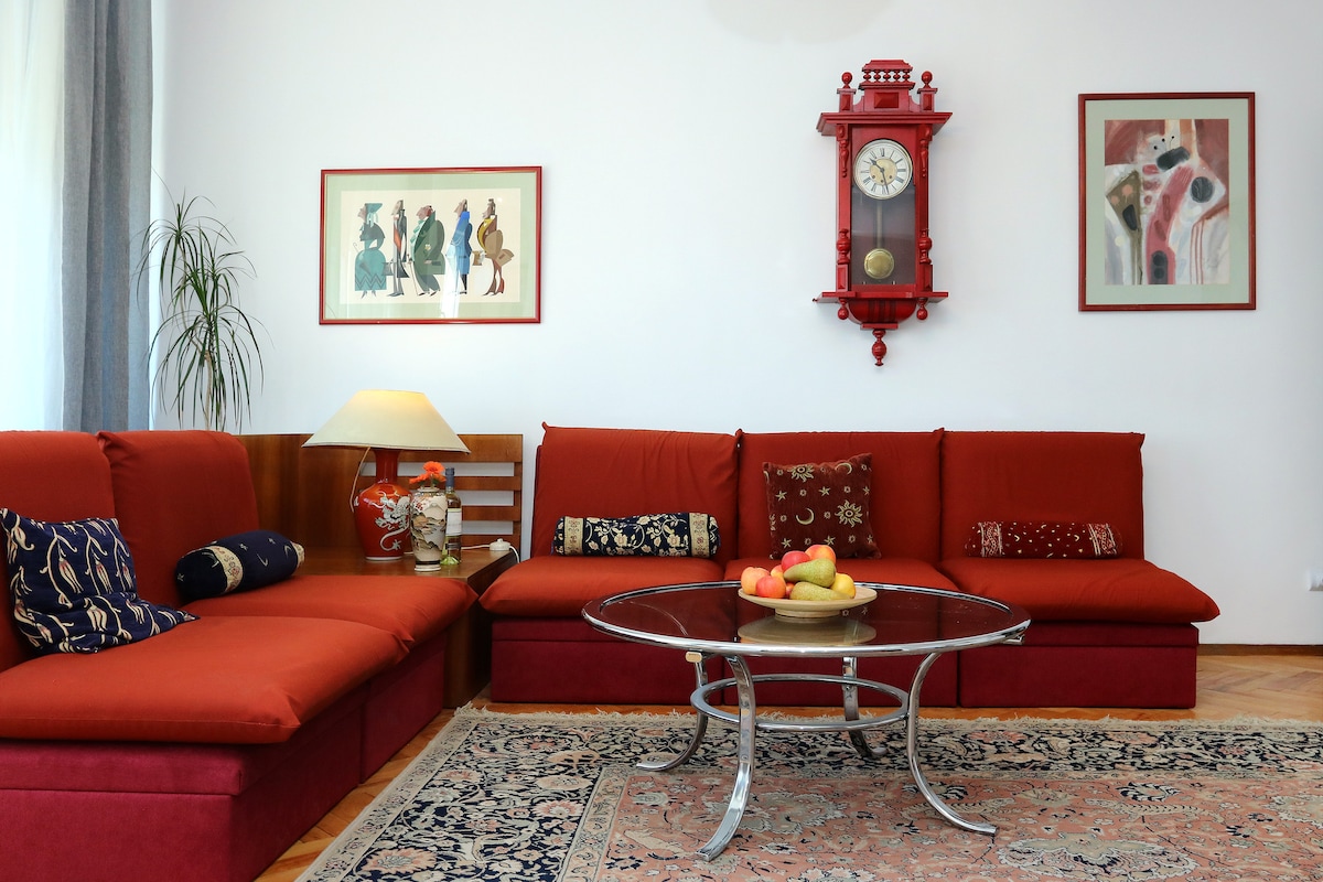 A bright living room features a red sectional couch adorned with various cushions. A round glass coffee table sits in the center, accompanied by a bowl of fresh fruit. Artwork and a vintage clock are displayed on the white walls, complementing the inviting atmosphere.