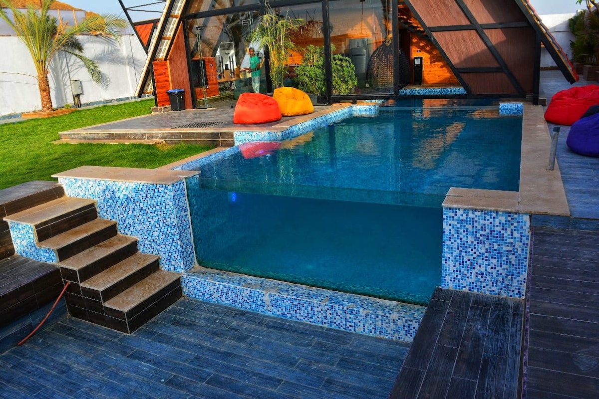 A swimming pool featuring an integrated area with a clear view, surrounded by patterned tiles. Colorful bean bags are placed along the pool's edge, and a wooden deck leads to steps descending into the water. Lush greenery can be seen in the background.