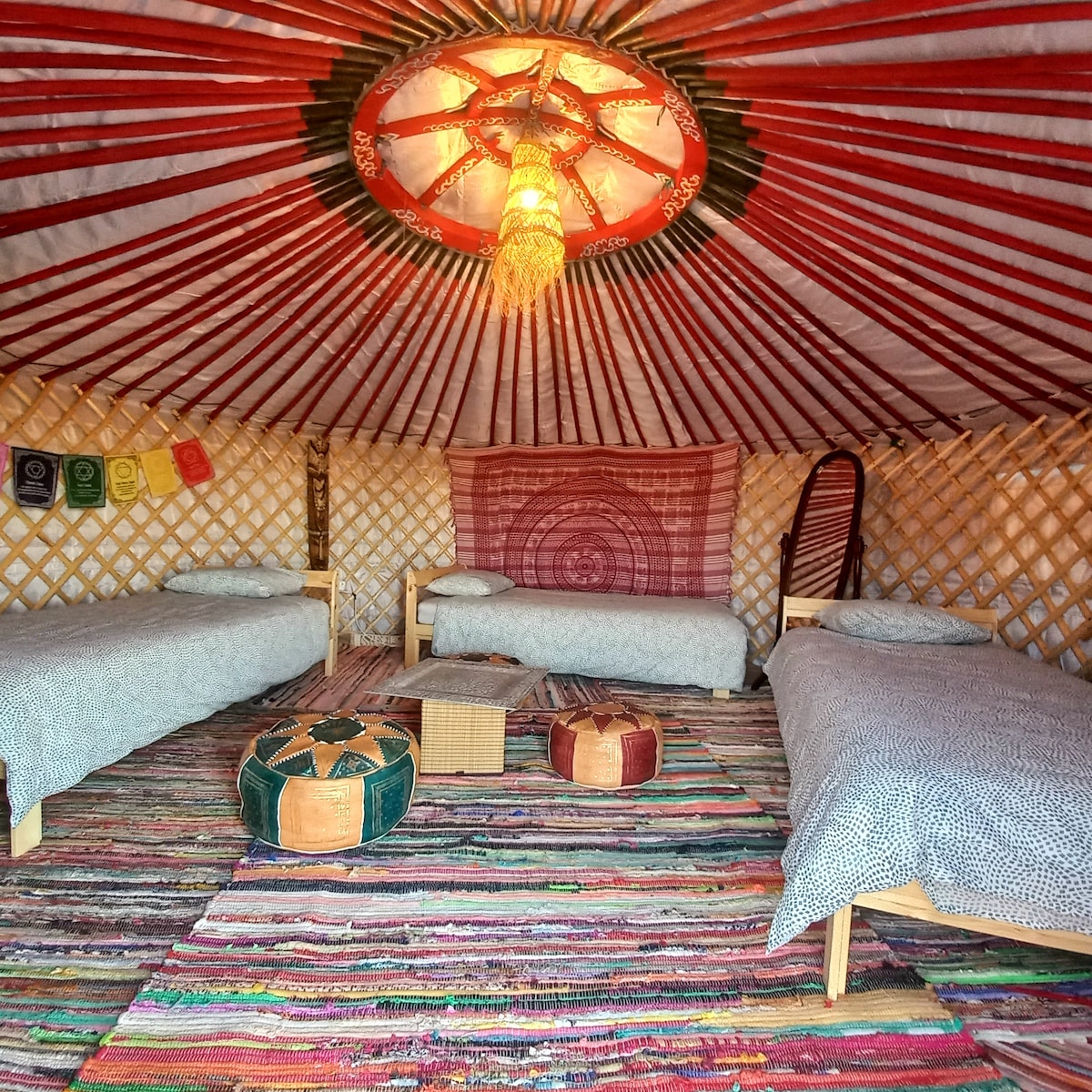 A spacious interior of a traditional yurt is shown with three beds arranged neatly. The colorful patterned ceiling adds warmth, complemented by woven rugs on the floor. Decorative elements, including lanterns and hanging fabrics, enhance the unique design of the space.