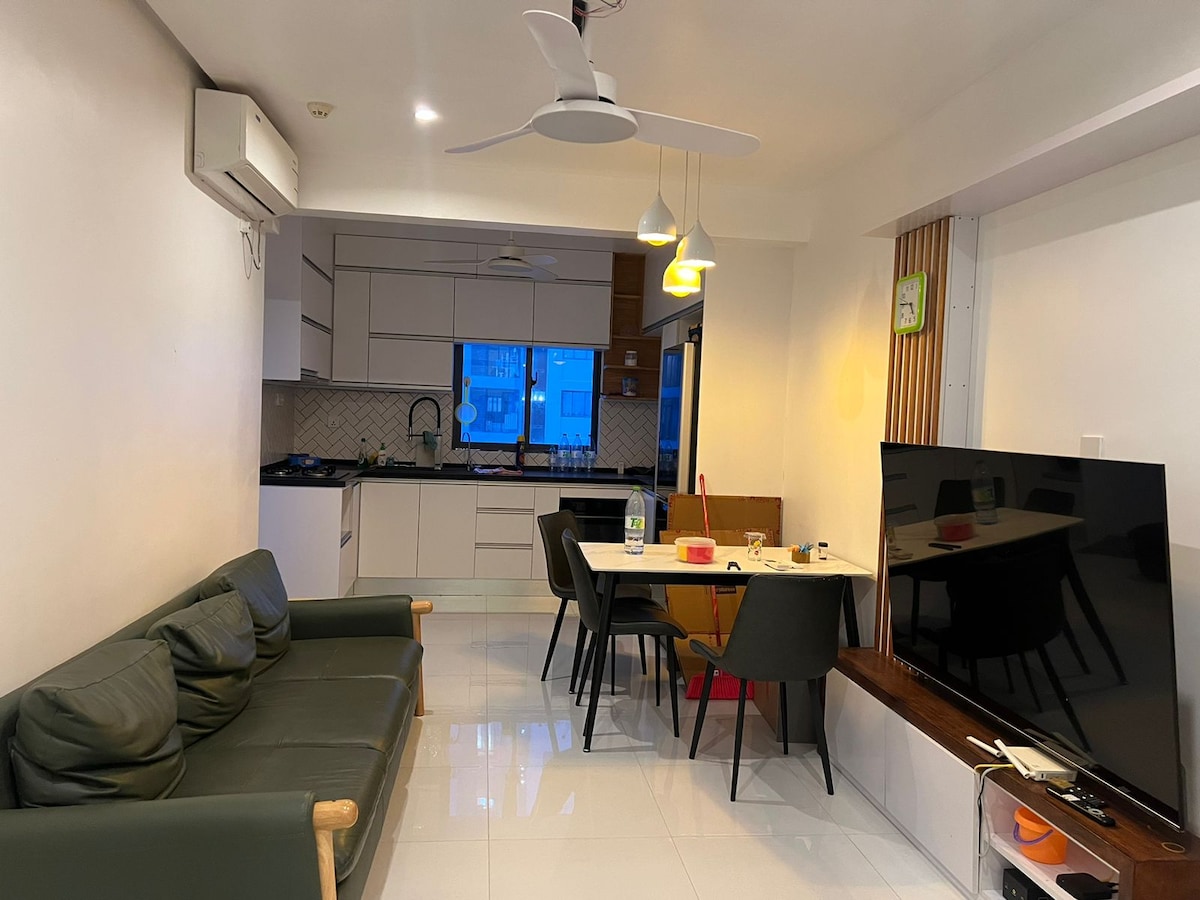 An open-plan living and dining area features a modern kitchen with sleek cabinetry and a dining table surrounded by several chairs. A comfortable sofa is positioned across from a large television, and natural light enters from a nearby window.