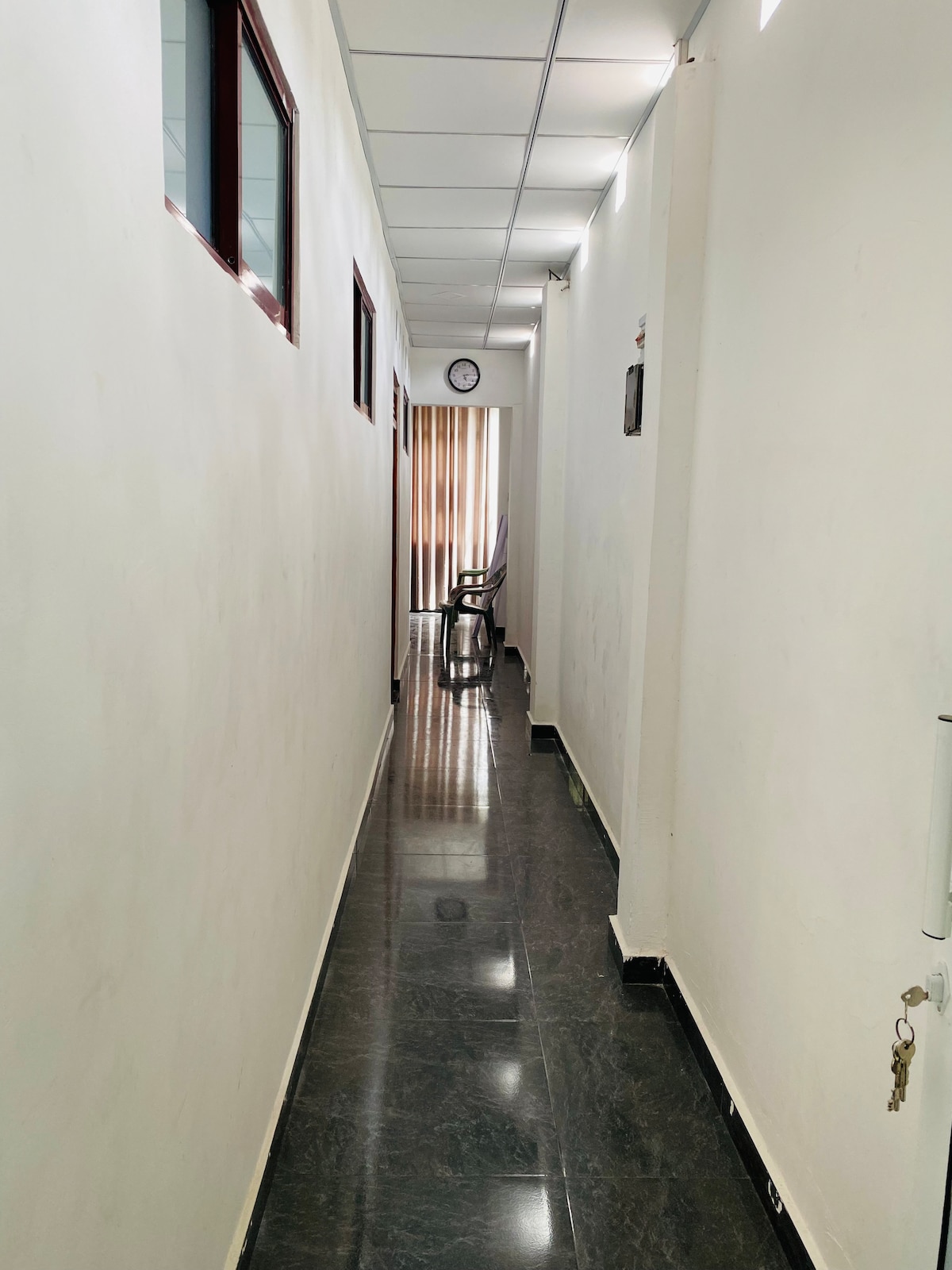 A narrow hallway is lined with light-colored walls and features a glossy dark floor. Natural light enters through windows along one side, illuminating the space. A clock is mounted on the wall, and a chair is positioned at the far end.