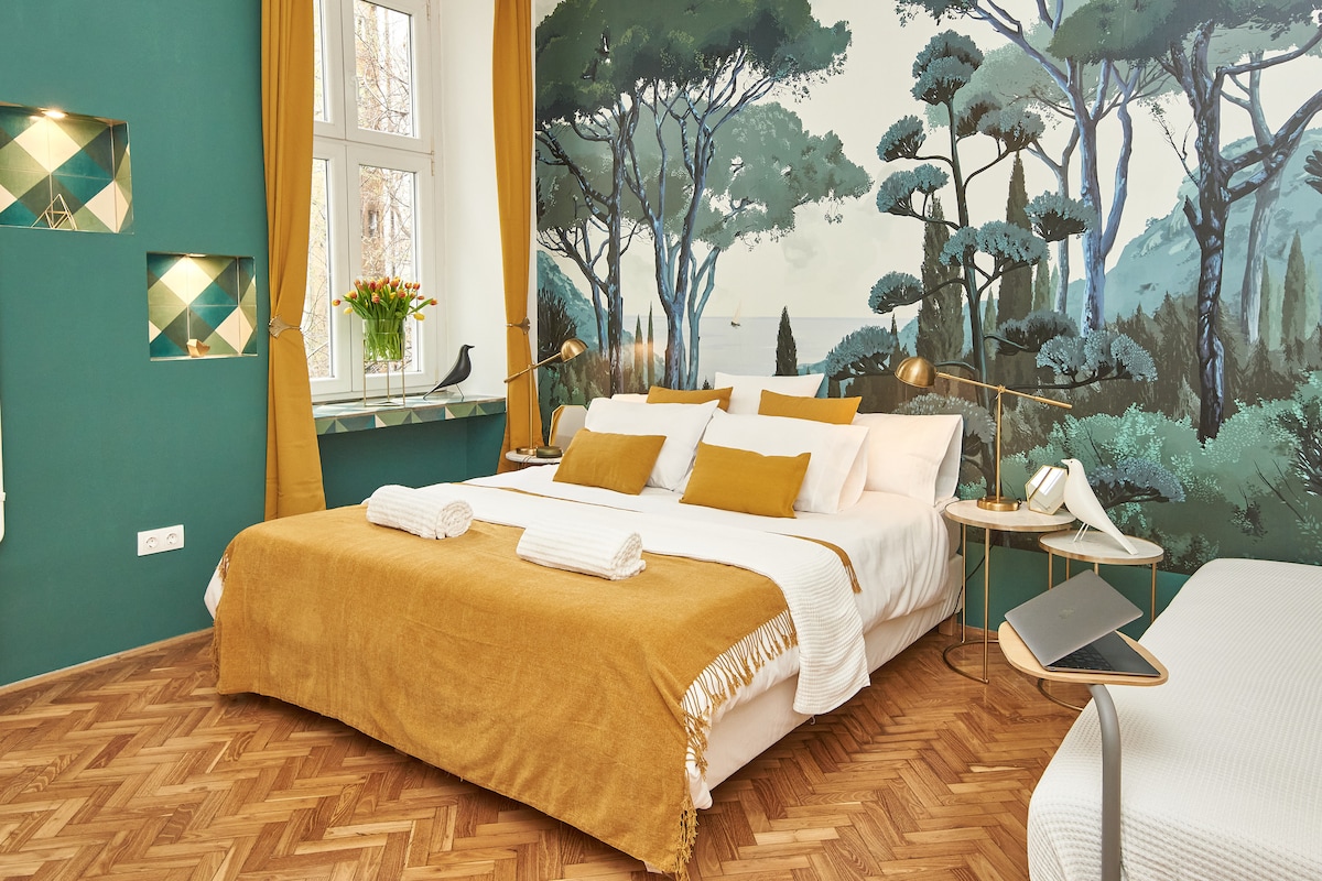 A spacious bedroom features a large bed adorned with a soft mustard-colored throw and bright white towels. A mural of trees creates a serene backdrop, while decorative fixtures and a bedside table complement the stylish decor. Natural light filters through large windows covered with golden curtains.