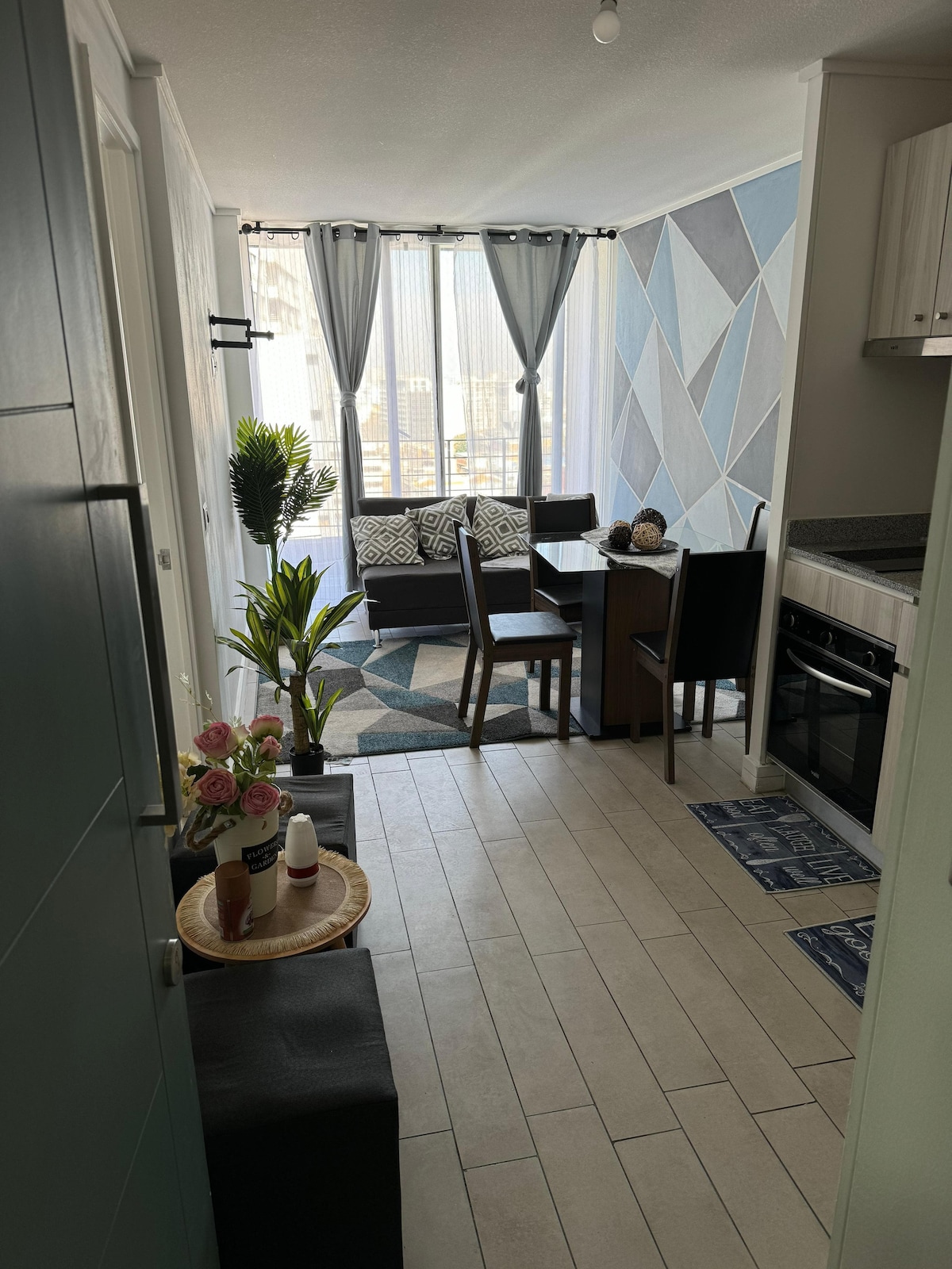 A modern living space is presented, featuring a dining area with a table and chairs, complemented by a cozy sofa. Large windows allow natural light to illuminate the room, while decorative plants and geometric wall designs enhance the aesthetic. A neutral color palette softens the atmosphere.