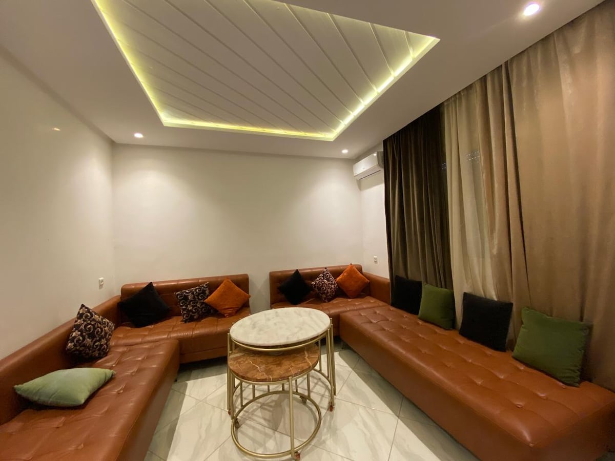A modern living area is presented with L-shaped leather seating positioned around a circular marble coffee table. The walls are adorned with neutral tones, and soft ambient lighting highlights the textured ceiling. Decorative pillows in various colors add subtle contrast to the space.