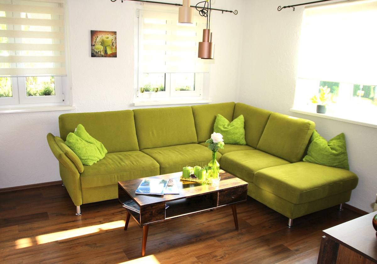 A bright living room features a green sectional sofa adorned with matching cushions, creating a comfortable seating area. A wooden coffee table is centered on the floor, with decorative items and magazines displayed. Natural light filters through two windows dressed with light blinds.
