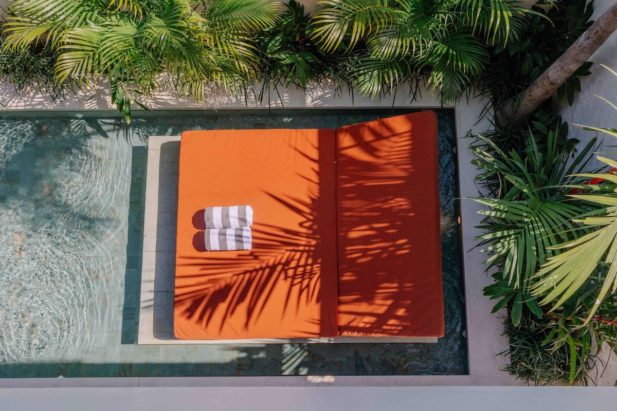A double sunbed is situated beside a refreshing pool, surrounded by lush tropical greenery. Two patterned towels rest on the sunbed, and shadows from palm leaves create an inviting pattern on the surface.