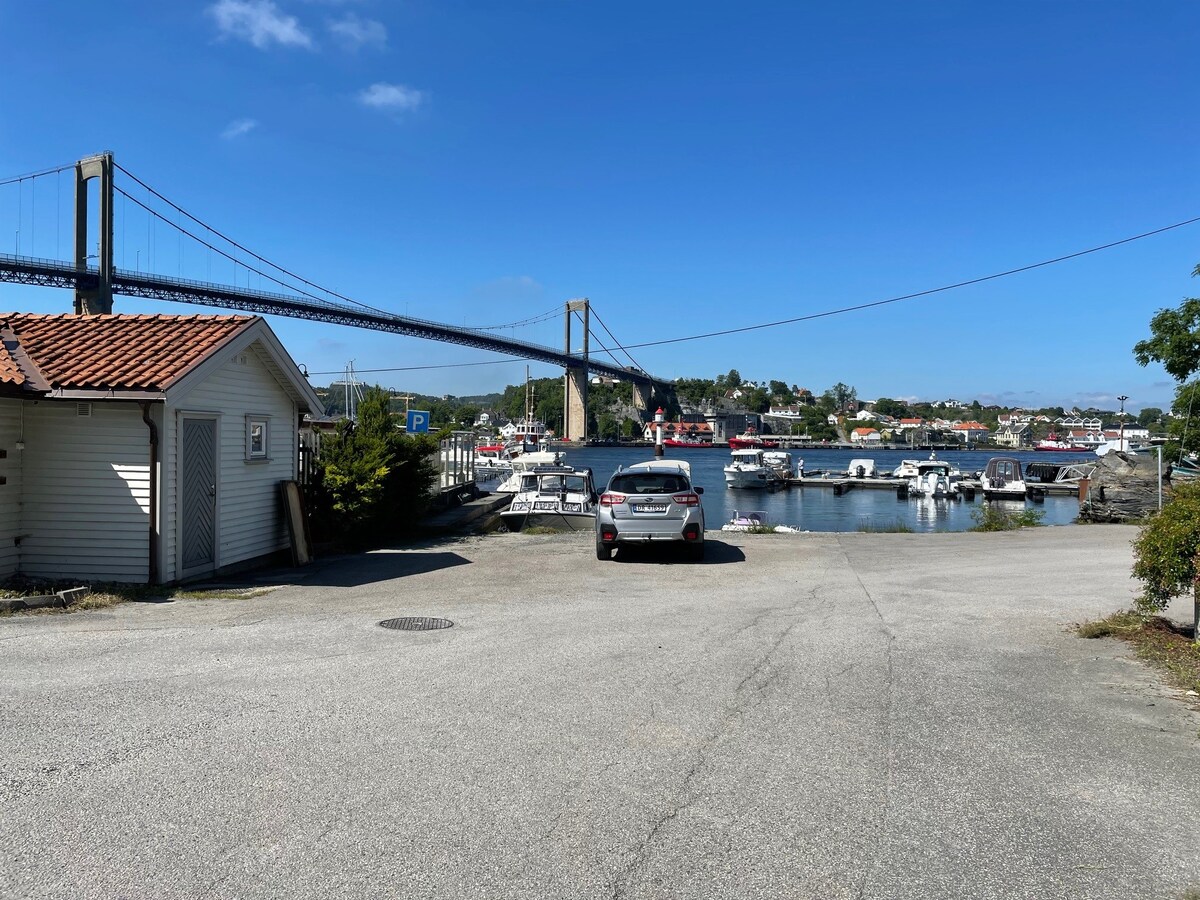 A scenic view showcasing a marina filled with boats and a bridge in the background. The image captures a clear blue sky with few clouds, while a parked car is positioned on a paved area near the water, providing a glimpse of the nearby village.