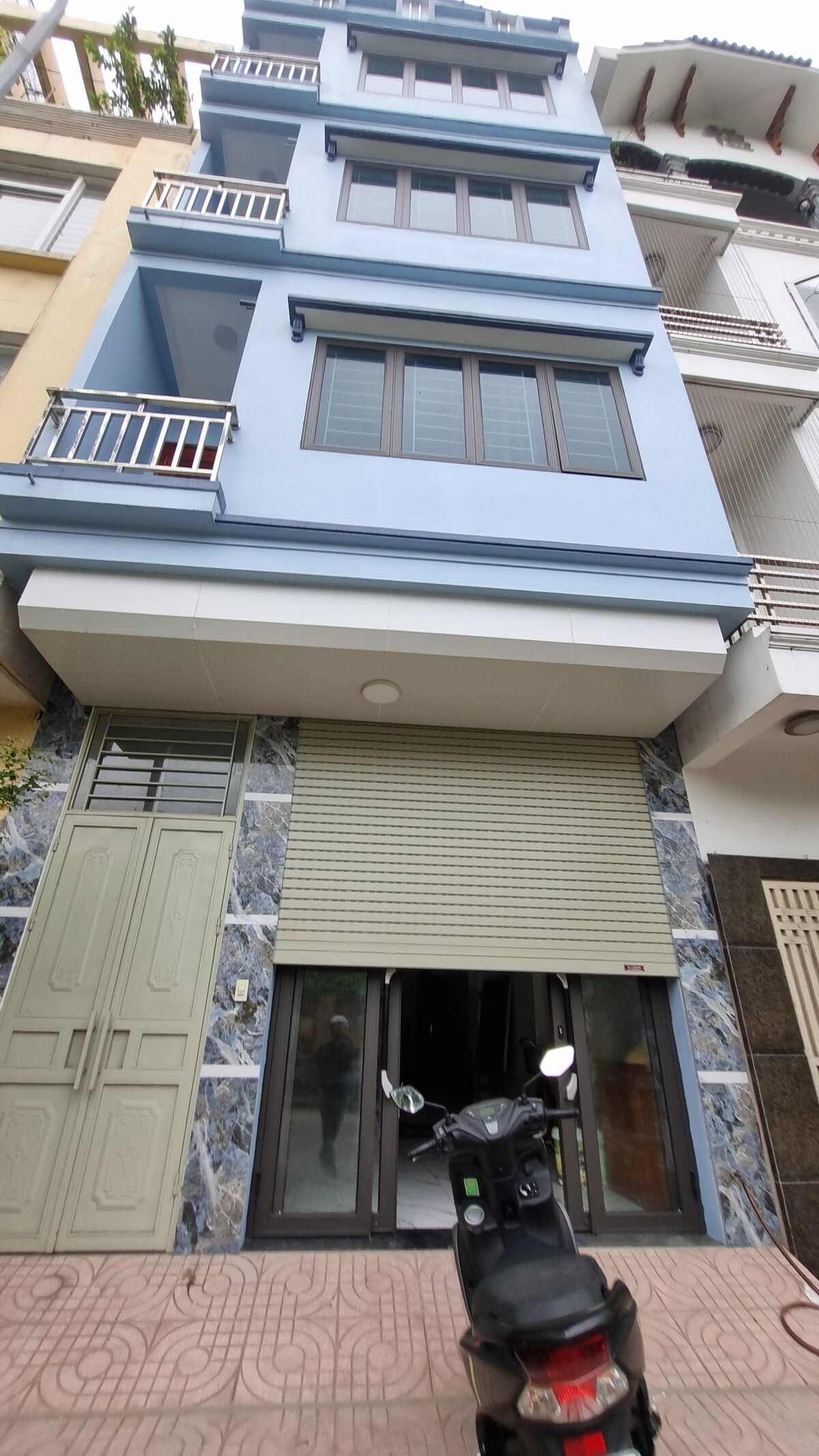 A three-story building with a light blue exterior features prominent balconies on the upper floors. The ground level showcases a large glass entrance, partially obscured by a rolled-up security shutter. A scooter is parked nearby on the paved walkway.