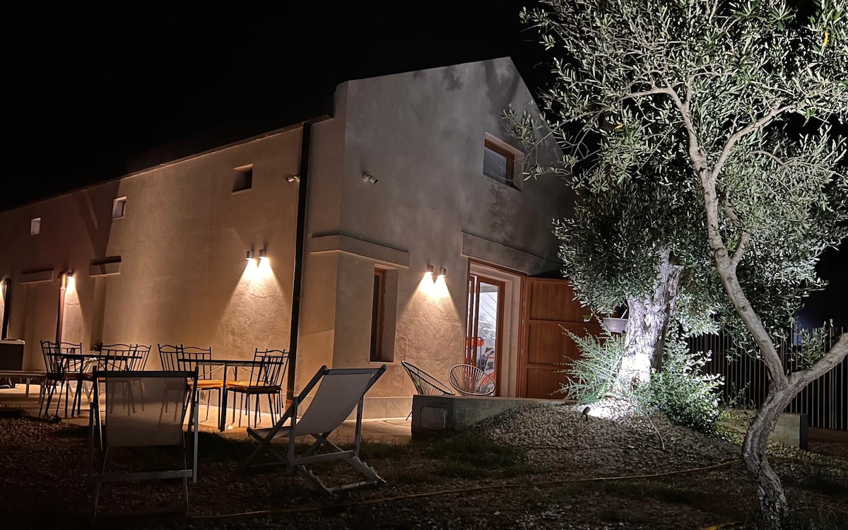The exterior of a modern loft is illuminated at night, featuring a warm entrance door and several outdoor dining tables and chairs. An ancient olive tree stands nearby, adding to the serene atmosphere of the surrounding landscape.