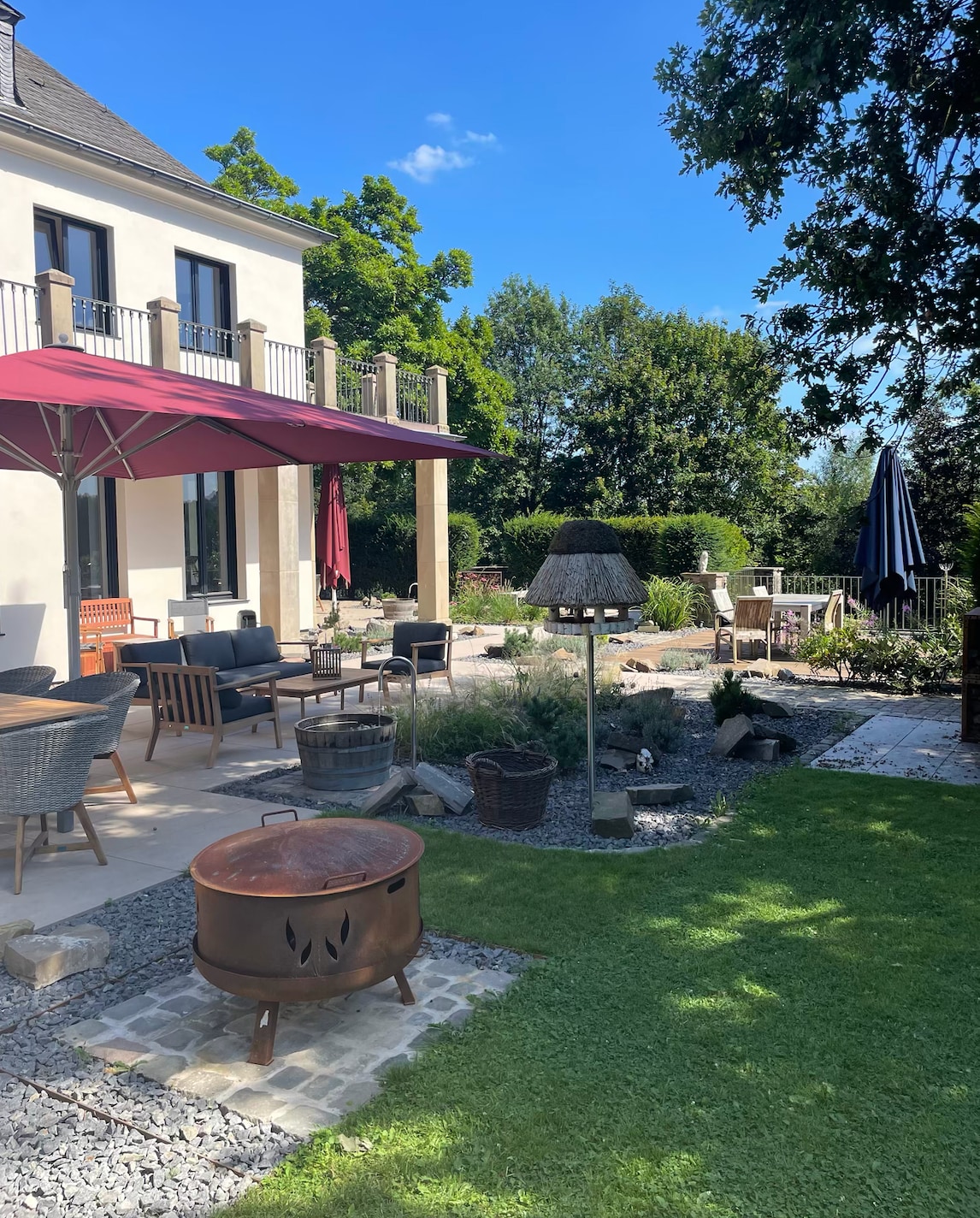 An outdoor area features a patio with seating arrangements, including stylish armchairs and a table. A large umbrella shades the space, while greenery surrounds a decorative fire pit. Soft lighting from a standing lamp enhances the inviting ambiance of the garden.