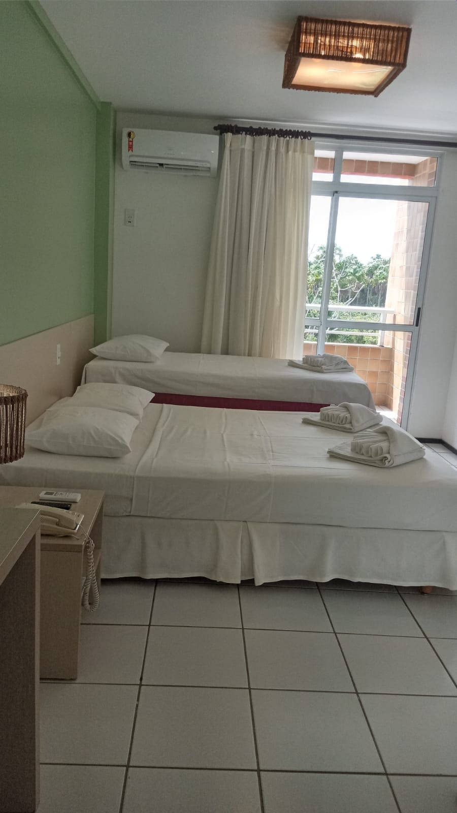 A bright and airy room features two beds with white linens, one being a spacious double bed alongside a single bed. Natural light floods the area through a large window, with sheer curtains softly diffusing the light. A small workspace and phone are visible.