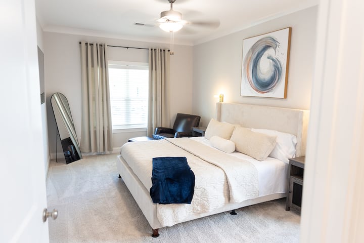 Dormitorio principal en la planta baja con TV Roku de 50 pulgadas, cortinas opacas, espejo de longitud completa, portaequipajes y silla de cuero con otomana.