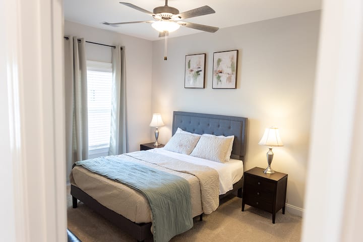 Cama tamaño queen con mesitas de noche, lámparas, cortinas opacas, arte de pared minimalista y otomanas