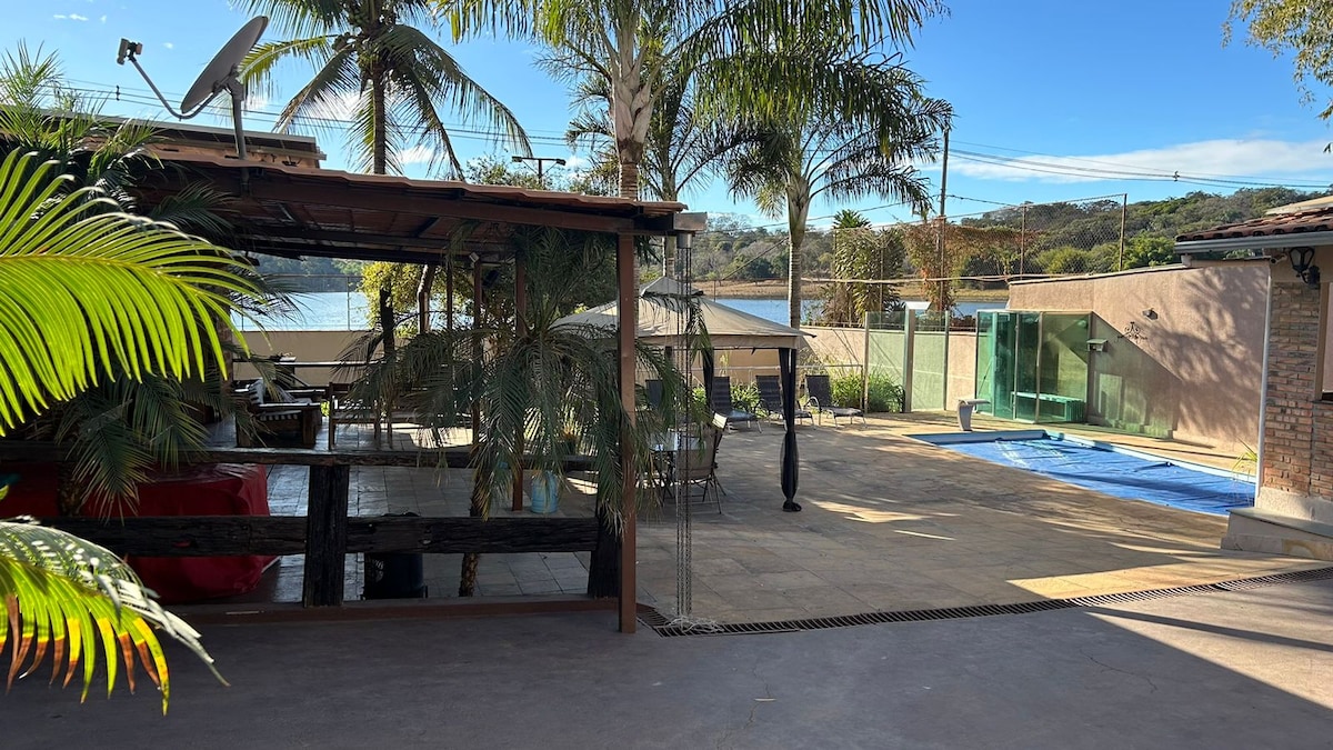 A spacious outdoor area is surrounded by palm trees and features a serene view of the lagoon. Comfortable seating options are arranged beneath a shelter, while a pool is visible nearby, enhancing the tranquil outdoor atmosphere.