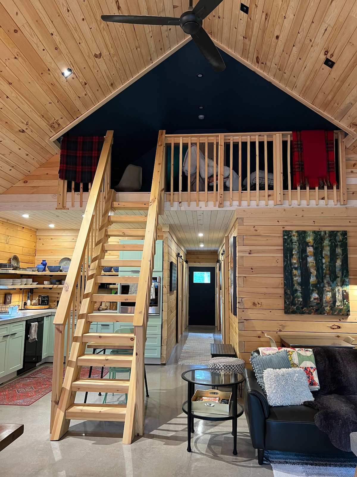 An open living space features wood-paneled walls and a high vaulted ceiling. A wooden staircase leads to an upper loft area lined with a balcony railing. A cozy seating arrangement is visible, complemented by a small coffee table and decor on nearby walls.
