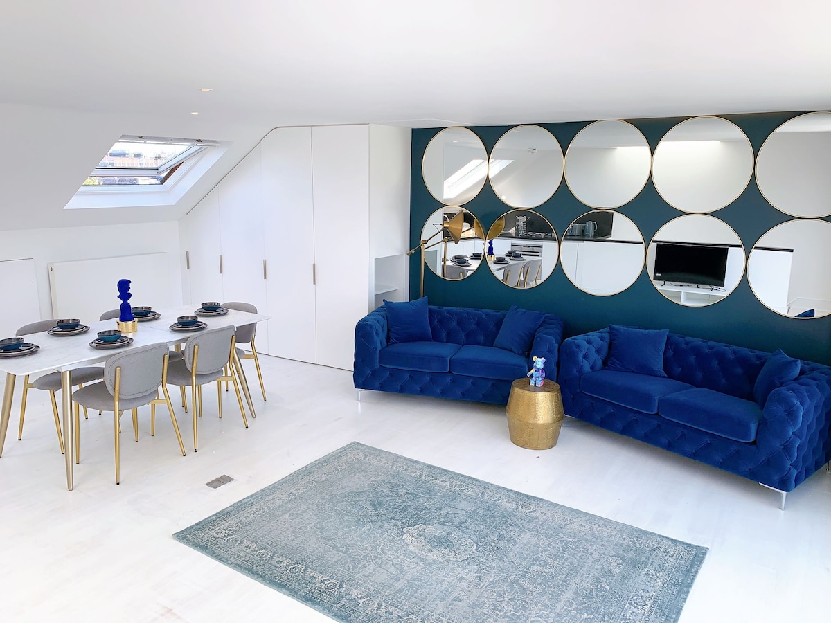 A contemporary living area is highlighted by two plush blue sofas and a stylish dining table set for six. A series of round mirrors reflects light, enhancing the space's brightness. A soft area rug adds texture to the light-toned flooring beneath.