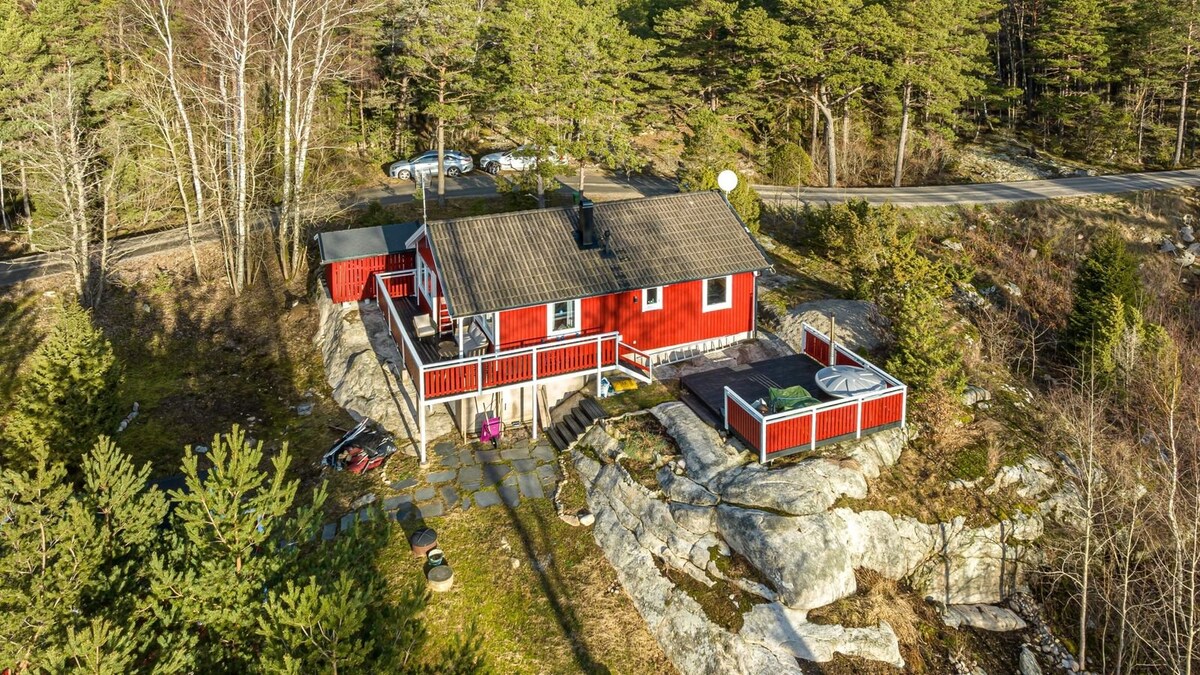 The image depicts a red cottage situated amidst a forested landscape. It features a spacious deck at the front, surrounded by natural stone and trees. A hot tub is visible on the deck, while additional outdoor space includes a stone pathway and a view of the surrounding greenery.