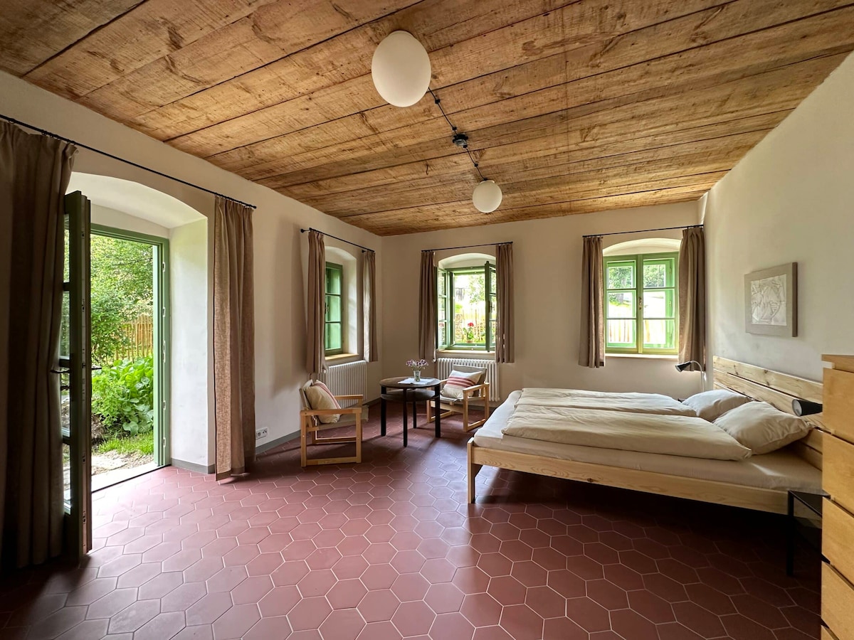 A spacious bedroom features a light wood bed with neatly arranged bedding. Natural light enters through two windows and a doorway leading to the garden. Two chairs and a small table are situated near the entrance, creating a cozy seating area.
