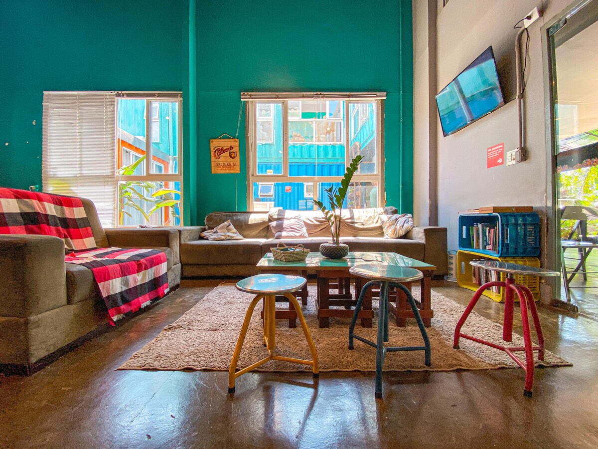 A communal lounge area is featured, adorned with teal walls and natural light streaming through large windows. A sofa is complemented by a plaid throw, while a coffee table sits atop a textured rug. Colorful stools add vibrancy to the space, creating an inviting atmosphere.