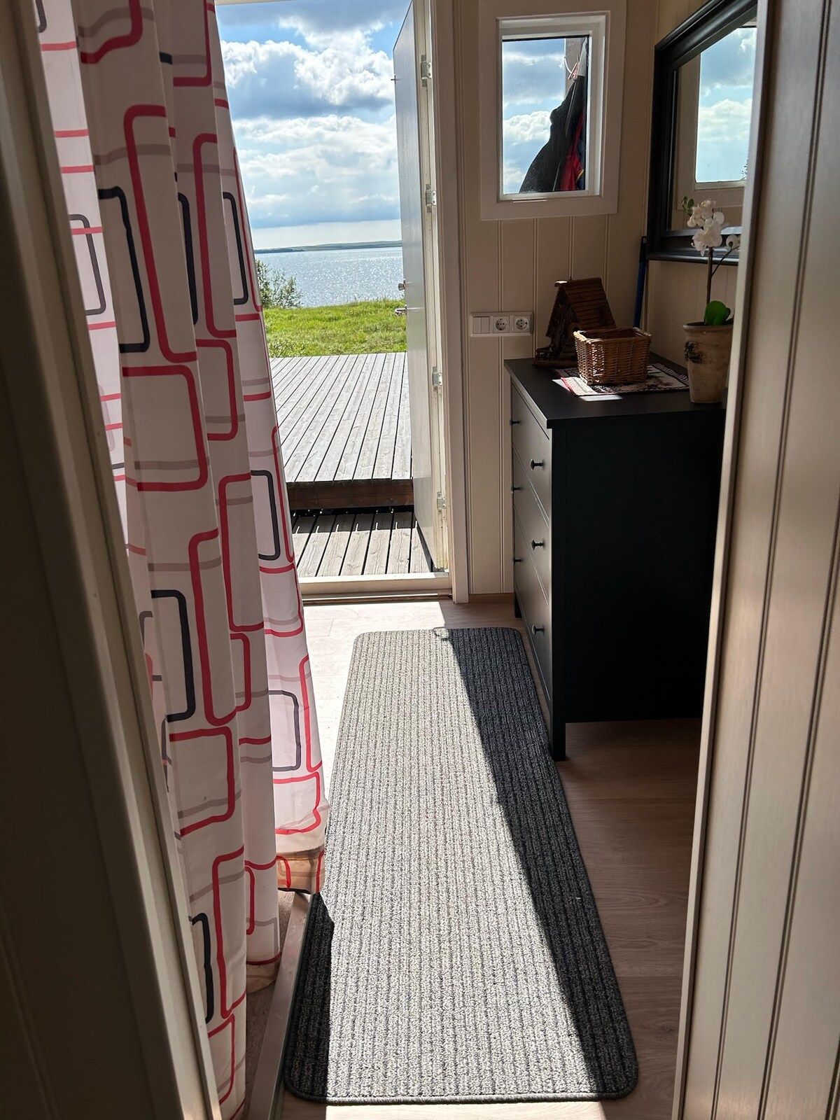 A welcoming entryway is seen leading to a deck with a lake view. Natural light brightens the area through a partially open door. A textured rug spans the floor, while a dark cabinet and decorative items are positioned against the wall.