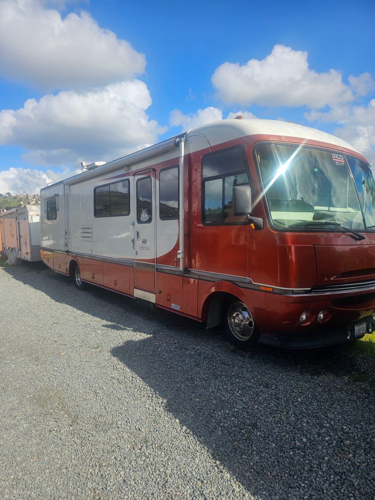 A vintage motorhome is parked on a gravel driveway, showcasing a distinct red and white exterior. The vehicle's rounded shape and large windows offer a nostalgic charm, while the bright blue sky with scattered clouds creates a vibrant backdrop.