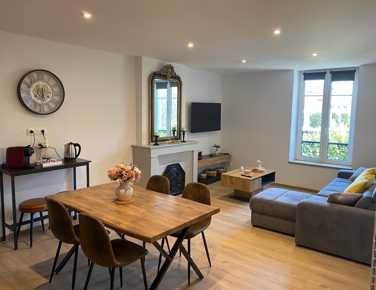 A spacious living area is featured, including a modern dining table with chairs and a comfortable sectional sofa. Natural light enters through windows, illuminating a decorative fireplace and a television mounted on the wall. A small coffee station is also visible.