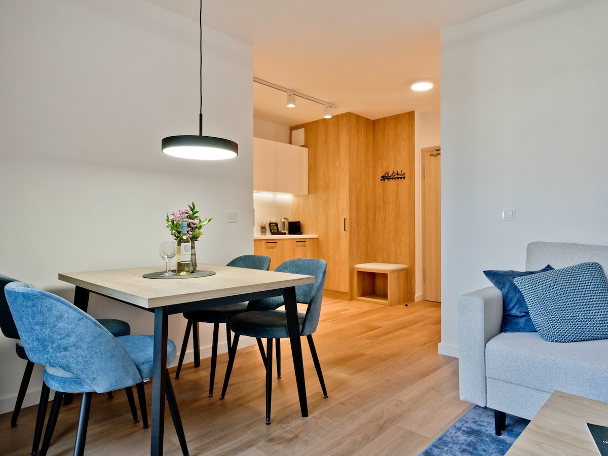 A dining area features a wooden table surrounded by four blue chairs. A small vase with flowers is displayed on the table. The background shows a well-equipped kitchen with white cabinets and a wooden accent wall, enhancing the cozy atmosphere.