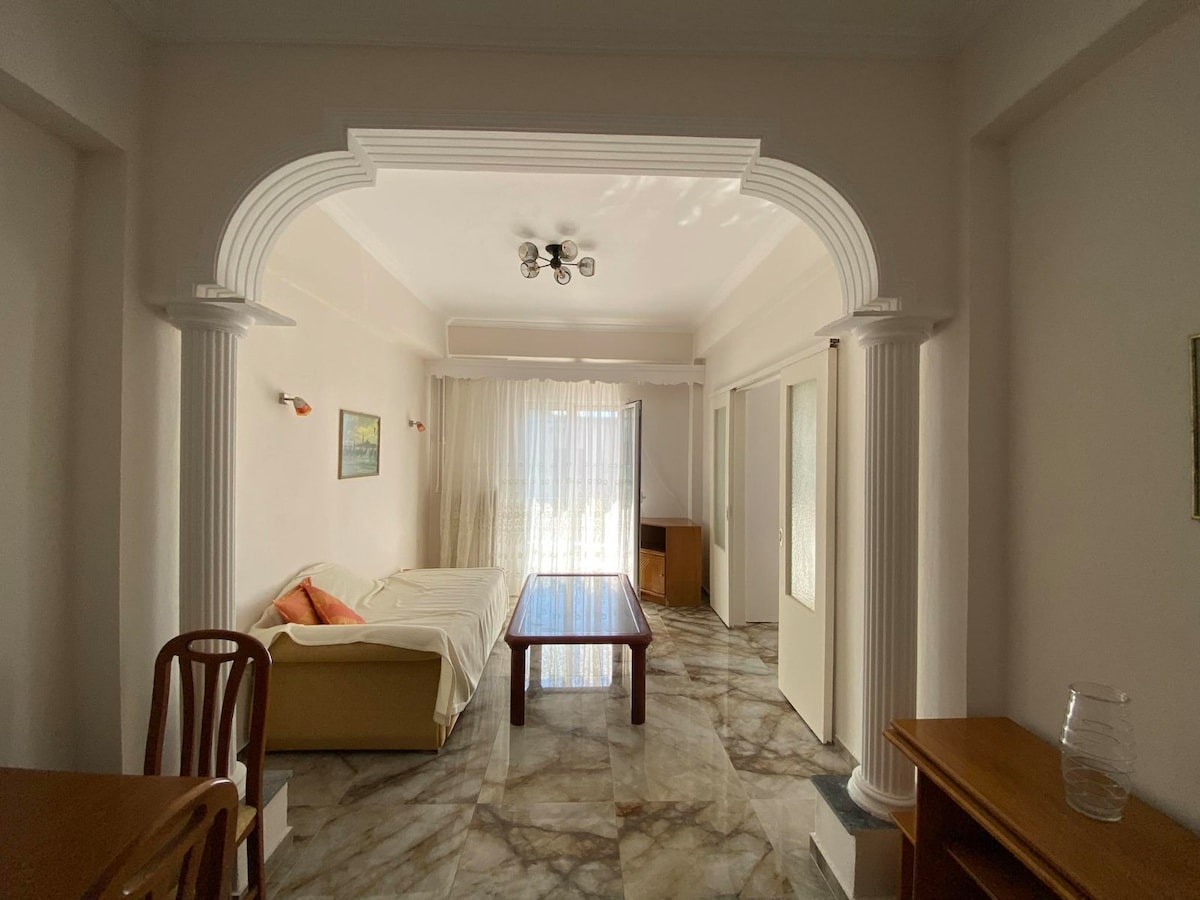 A spacious living area features natural light streaming through large windows, creating a bright atmosphere. A comfortable sofa and a wooden dining table are visible, along with light-colored walls and marble flooring that enhance the open layout. Decorative elements include a framed painting on the wall.