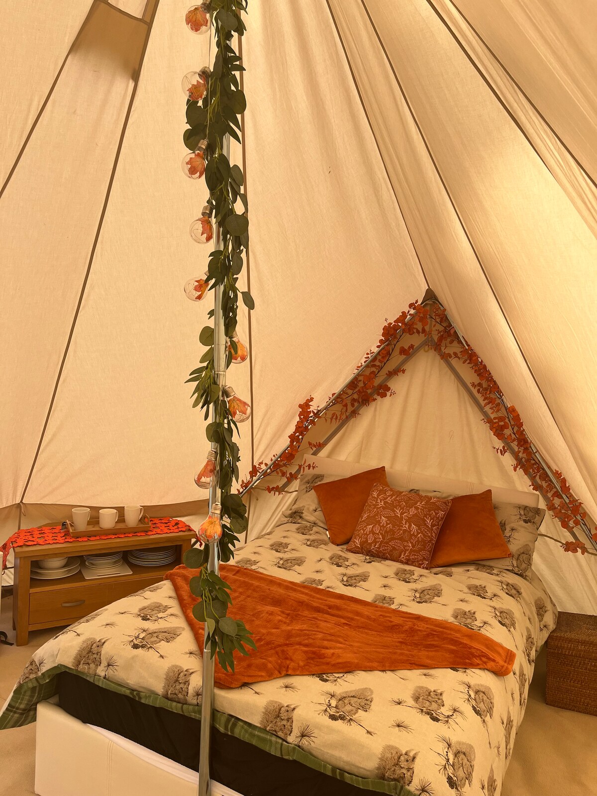 A cozy glamping tent features two real double beds adorned with decorative pillows and blankets. Strings of soft lights hang from the center pole, surrounded by autumn-themed decorations. A wooden table is visible in the background with kitchenware arranged neatly, creating a welcoming atmosphere.