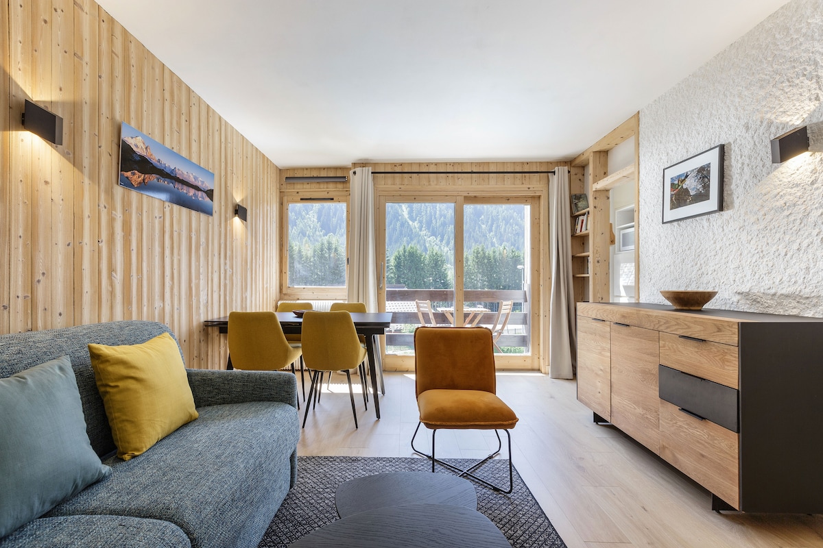A light-filled living area features a comfortable blue sofa with yellow pillows. A wooden dining table for four is visible, positioned near large windows offering views of the surrounding mountains. A wooden sideboard and a stylish chair complete the inviting space.
