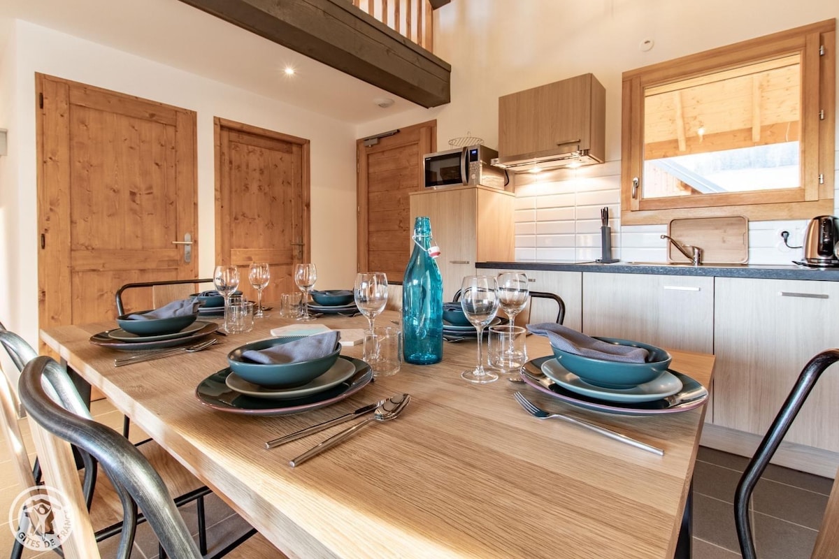 A dining area features a wooden table set for six, complete with plates, cutlery, and glassware. A blue glass vase is present in the center. Kitchen cabinetry is seen in the background, with a microwave and a sink visible. Large windows allow natural light to enhance the space.