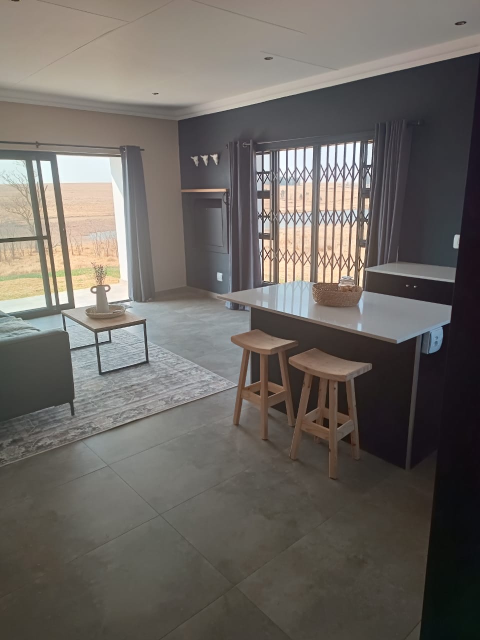 A contemporary living space is featured with a neutral color palette. Natural light pours in through large glass doors leading to an outdoor view. A small dining area is created with two wooden stools positioned at a sleek island countertop, enhancing the overall open feel.