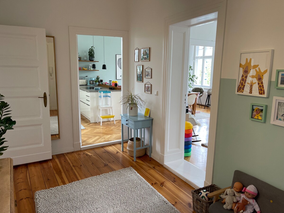 An entryway is presented with a textured rug and wooden flooring. A mirror and a small blue table are positioned beside a doorway leading to a spacious kitchen. Walls are adorned with framed art, and children's toys are visible in a corner, adding a playful touch.