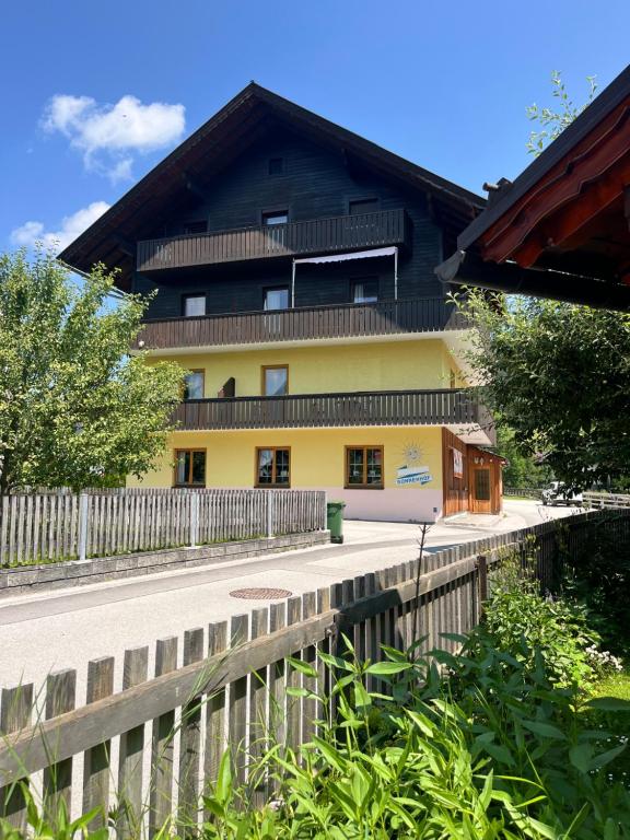 A multi-story building features a mix of dark wood and yellow exterior. Balconies are visible on the upper floors, and large windows allow natural light to enter. A well-maintained fence runs along the property, with greenery that enhances the outdoor space.