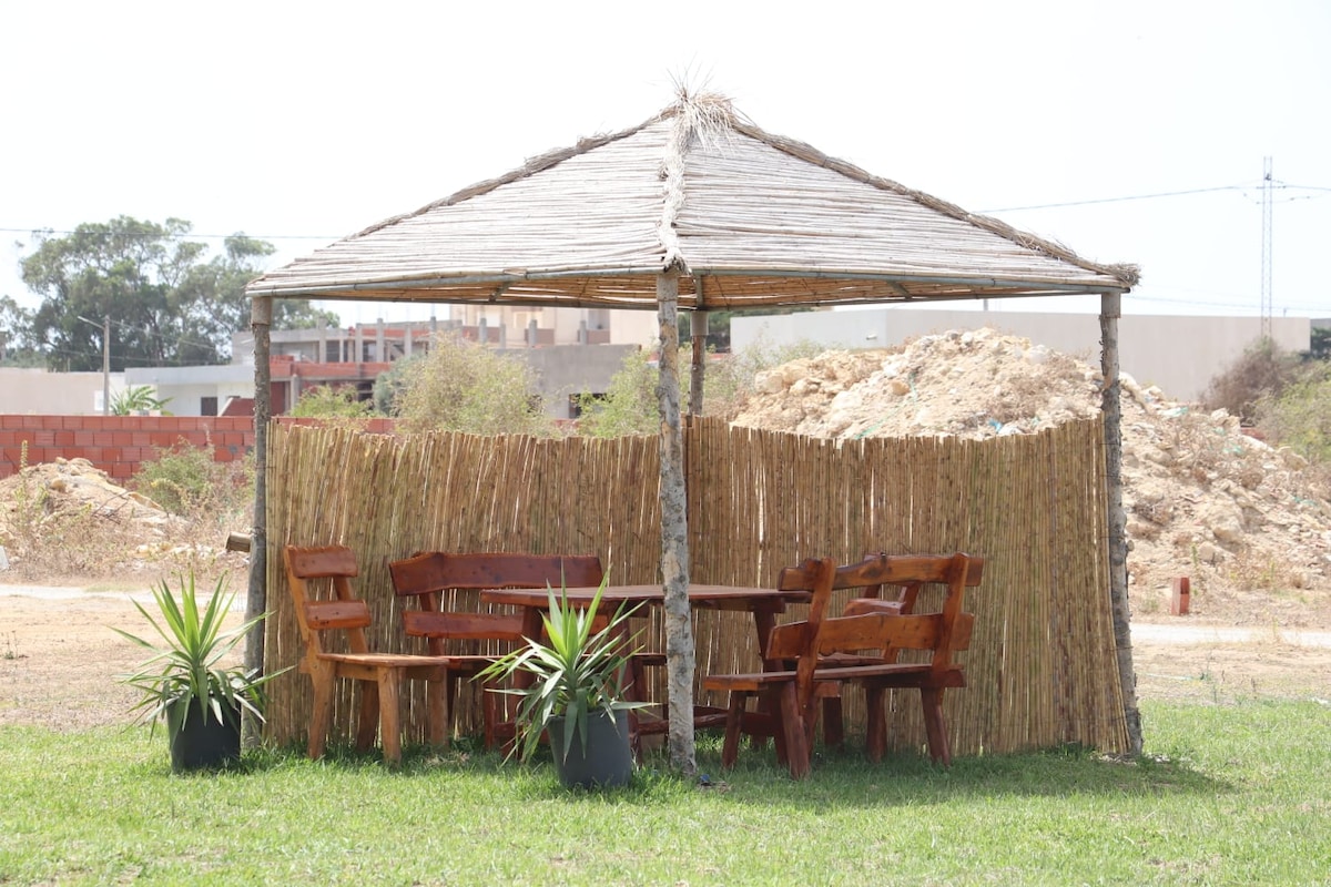 A shaded outdoor sitting area is surrounded by natural materials, featuring rustic wooden furniture, including a round table and several chairs. Potted plants are placed near the seating, and a thatched roof provides shelter. The backdrop includes sparse vegetation and distant structures.