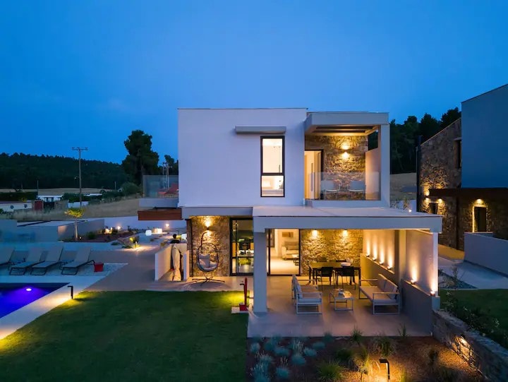 A modern villa exterior is illuminated under twilight, showcasing a private pool and patio area. Comfortable outdoor seating and a dining area are visible, set against a backdrop of natural landscaping. The architectural design features clean lines and large windows, enhancing the overall aesthetic.