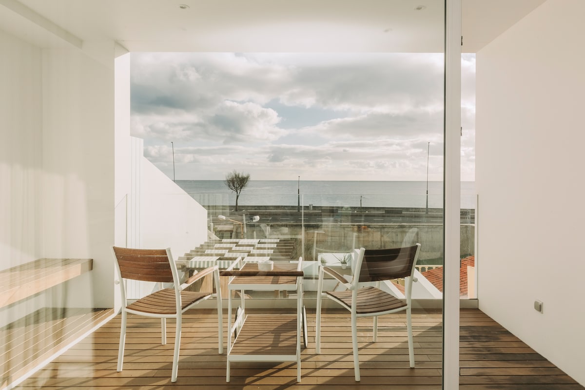 A modern terrace with wooden flooring offers a glimpse of the ocean. Two white chairs and a small table are positioned to enjoy the view, which includes a serene stretch of water under a cloudy sky.