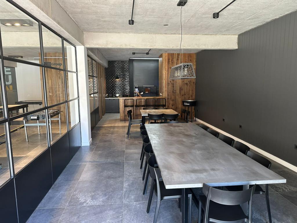 A spacious dining area is provided with a long, rectangular table, surrounded by black chairs. A modern pendant light hangs above the table. Large windows allow natural light to illuminate the room, complemented by wood and stone accents on the walls.