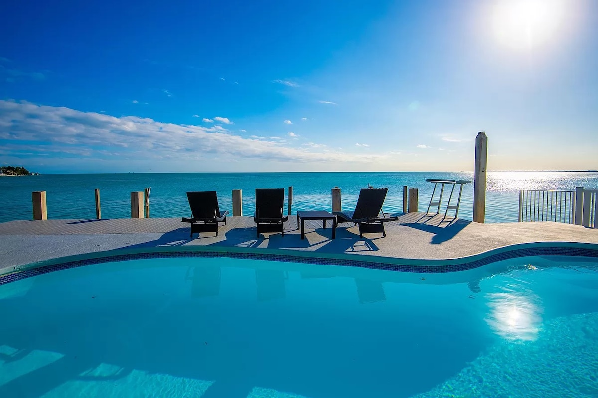 A private pool with clear blue water extends toward the ocean, surrounded by lounge chairs. The unobstructed ocean view showcases a bright sky, reflecting sunlight on the water's surface. A dock structure is partially visible, enhancing the outdoor living space.