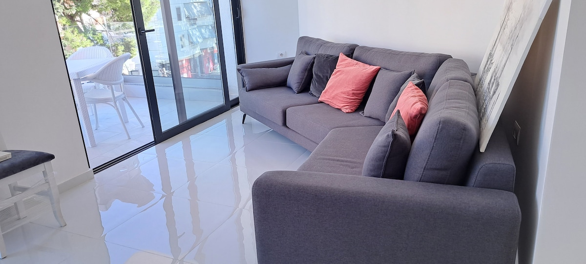 A modern living area is showcased with a gray sectional sofa adorned with coral accent pillows. Large glass doors provide a view of the outdoor space, while bright natural light reflects off the glossy white tile floor.