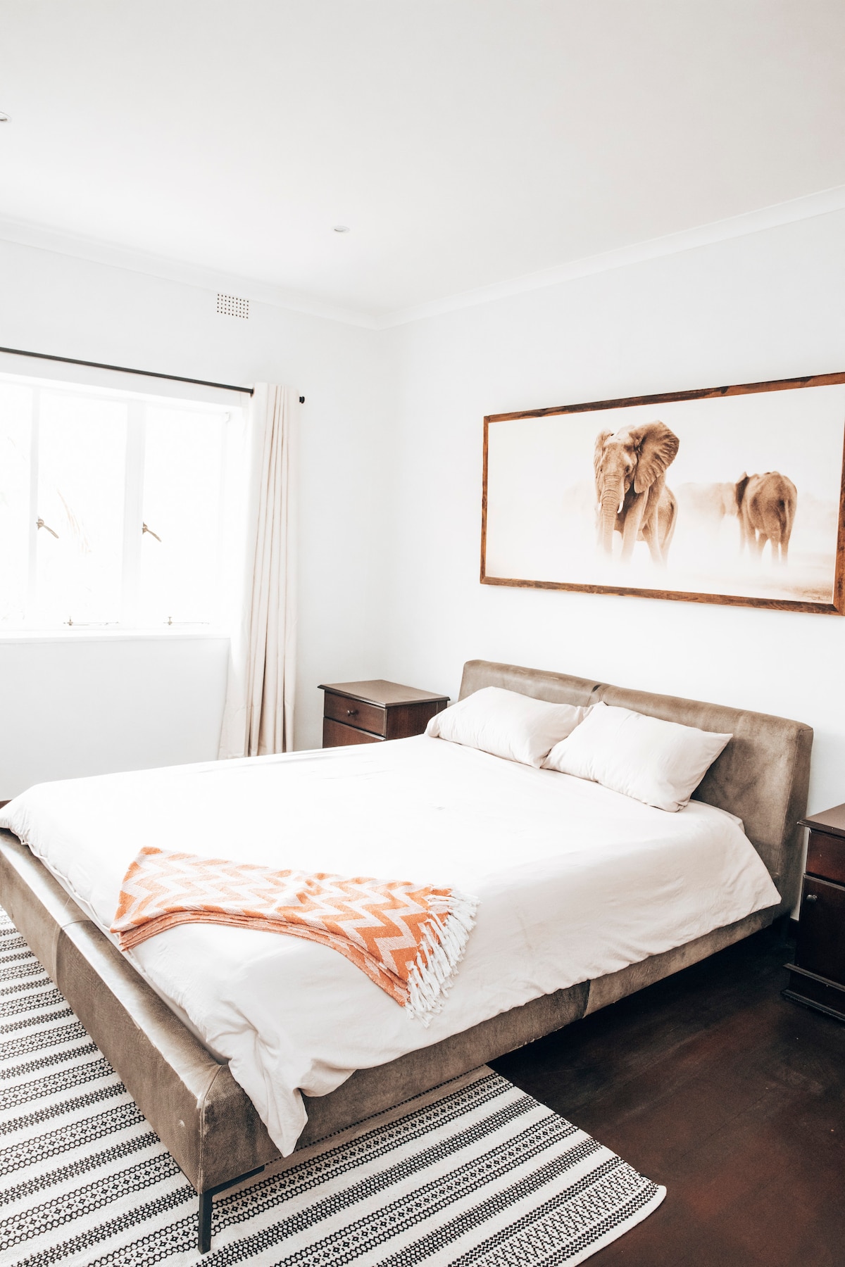 A light and airy bedroom features a spacious bed with neutral bedding, complemented by an orange patterned throw. Flanking the bed, two dark wood nightstands provide additional storage. A large window allows natural light to illuminate the room, while an elephant photo adorns the wall, adding a touch of character.