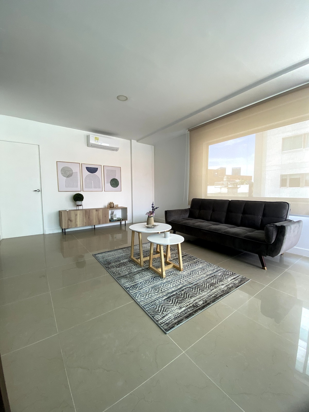 A modern living area is presented with a sleek black sofa, two small round tables, and a decorative rug. Wall art with minimalistic designs adorns one side, while natural light filters through a large window, creating a bright and airy ambiance.
