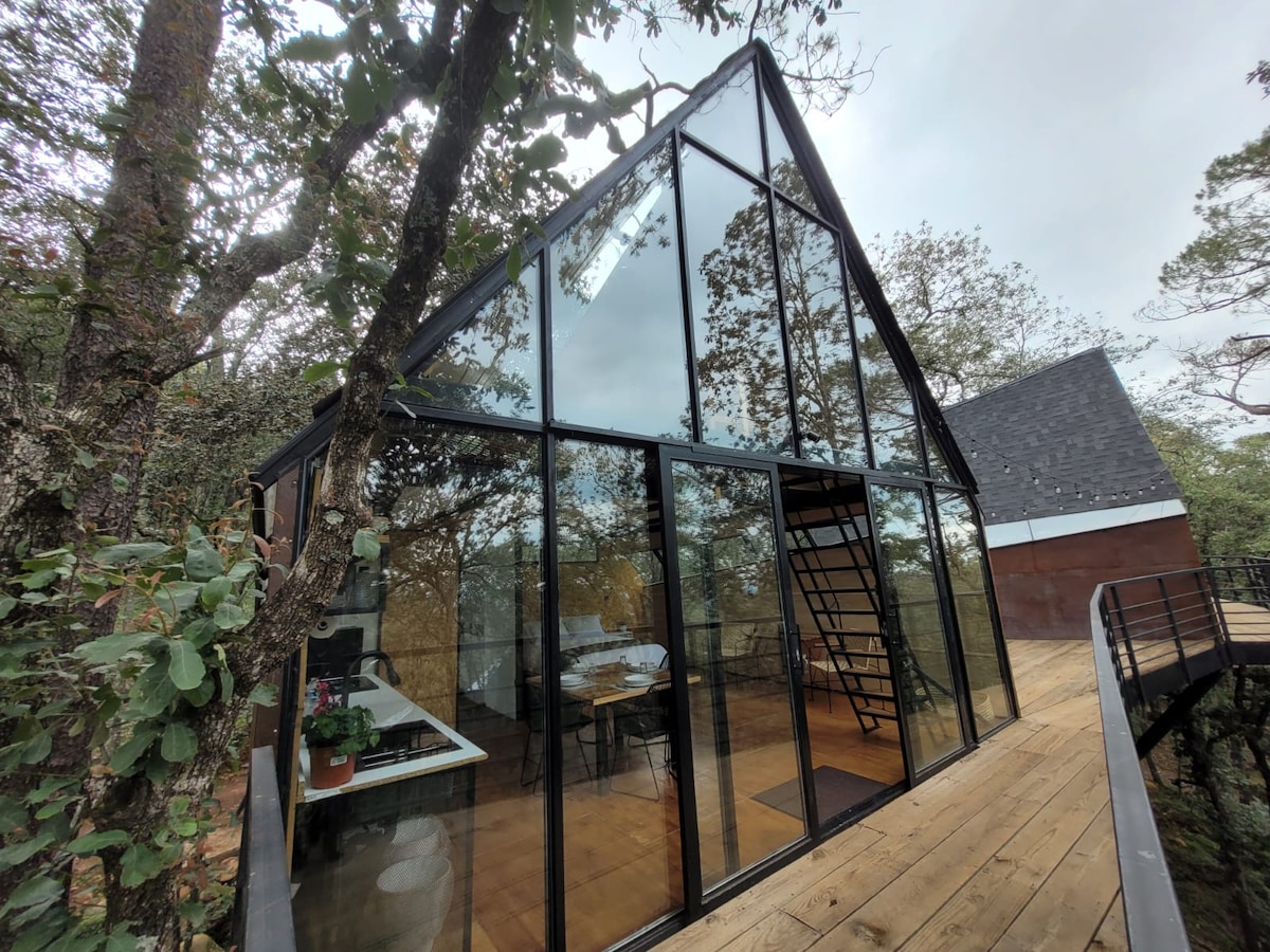 A modern elevated cabin features a striking glass facade, blending with the surrounding trees. A wooden deck leads to the entrance, showcasing a spacious interior filled with natural light. The geometric design is complemented by an inviting angle, emphasizing the connection to nature.