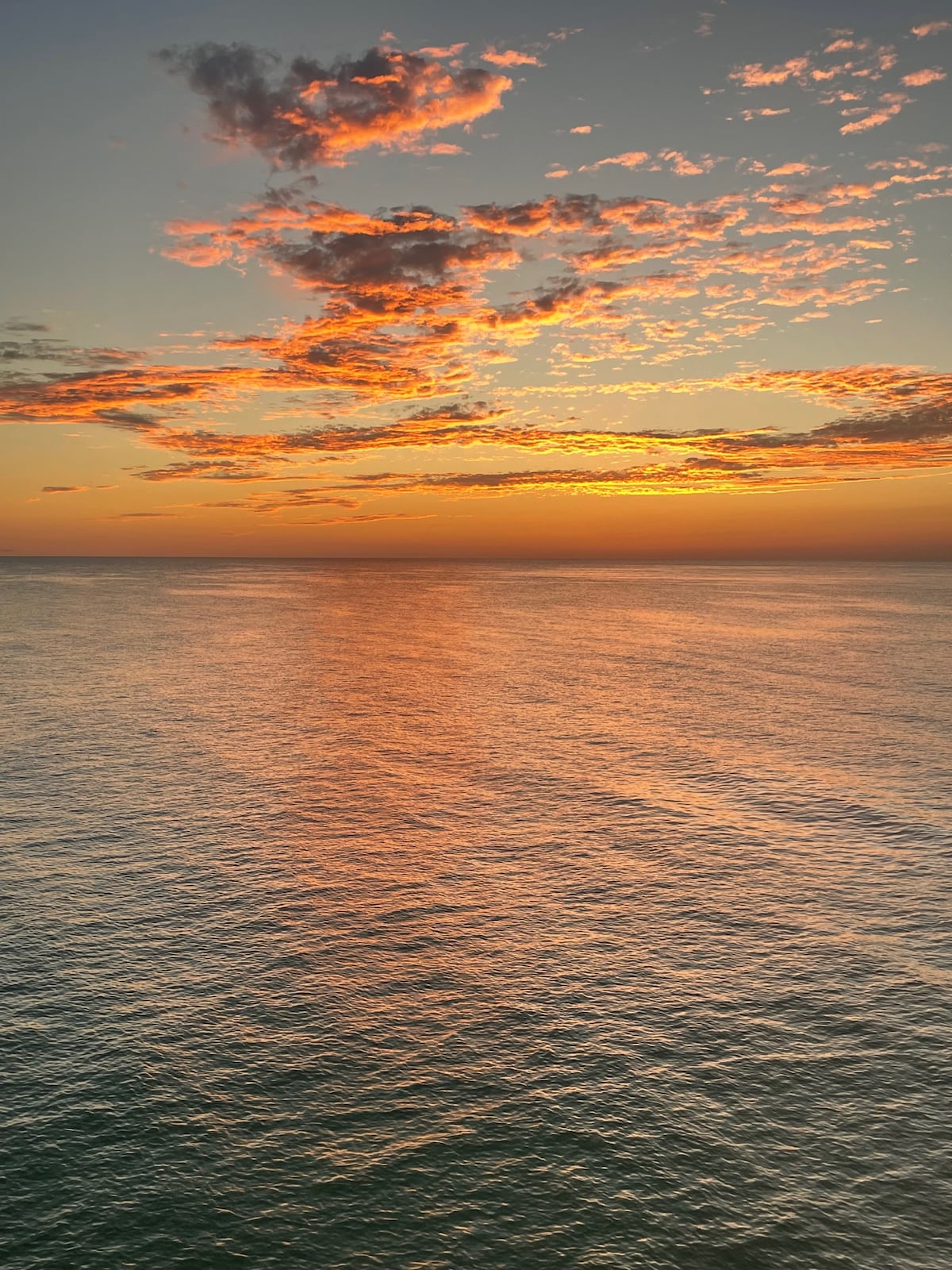 A vibrant sunset is depicted over calm ocean waters, with hues of orange and purple reflecting on the surface. Wispy clouds are visible, enhancing the serene atmosphere of the scene, which creates a tranquil backdrop for relaxation.