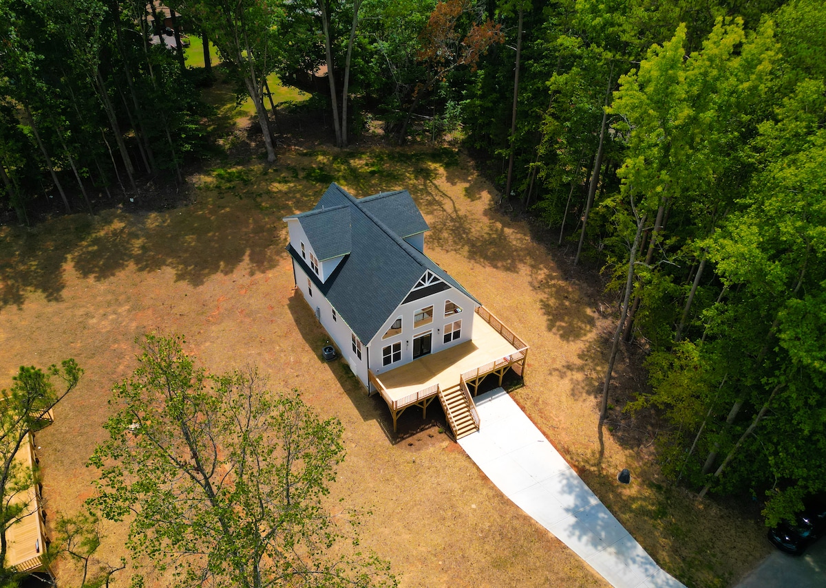 An aerial view of a modern house surrounded by trees in a serene outdoor setting. A spacious deck extends from the front, leading to a well-maintained driveway that provides clear access to the home.