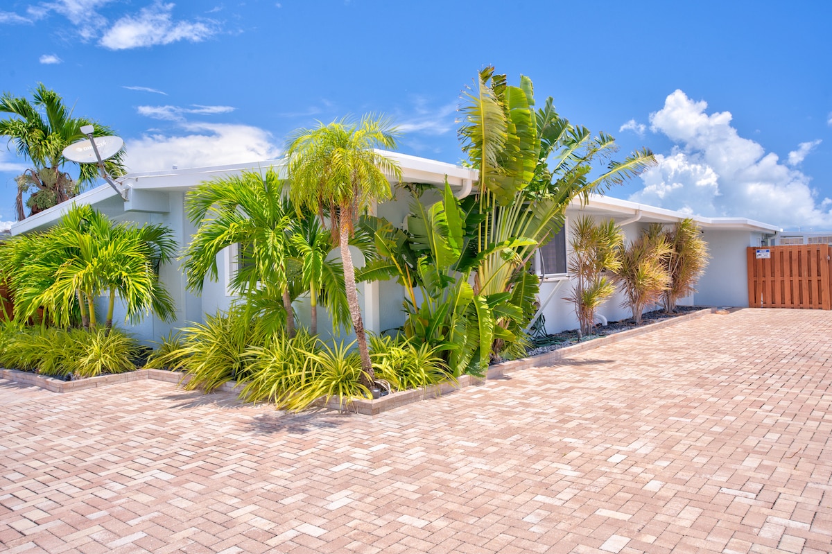 A welcoming exterior view of the property showcases a landscaped front yard with lush palm trees and tropical plants. A brick driveway leads to the entrance, complemented by a wooden privacy fence in the background under a clear blue sky.