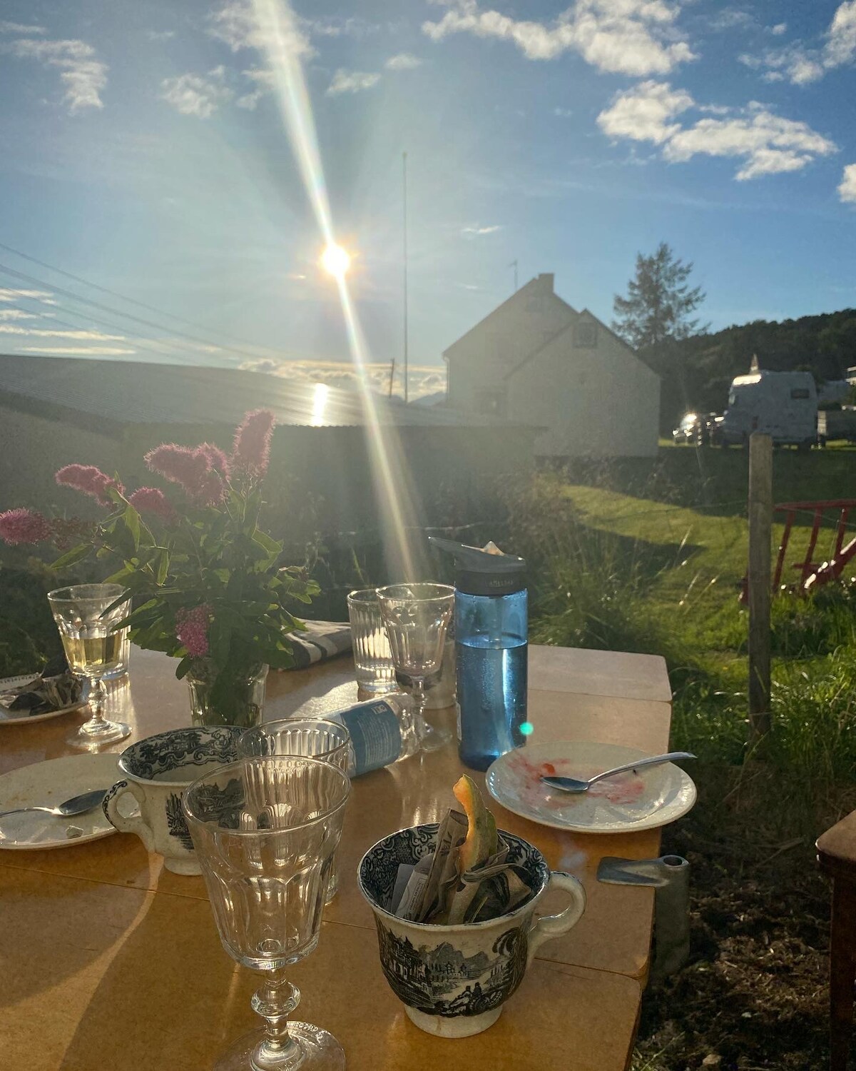 A table is set outdoors, adorned with glasses and a vase of flowers. The sun shines brightly in the background, casting a warm glow on the scene. A glimpse of a house and greenery can be seen in the distance, enhancing the tranquil outdoor setting.